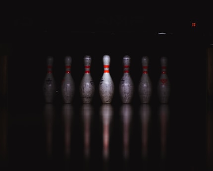 A row of seven bowling pins is aligned on a reflective surface, illuminated against a dark background. The pins have red stripes and show signs of wear and use.