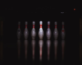 A row of seven bowling pins is aligned on a reflective surface, illuminated against a dark background. The pins have red stripes and show signs of wear and use.