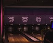 A dimly lit bowling alley features several lanes with bowling pins neatly arranged at the end. The scoring screens above the lanes display a stylized logo of a heart intertwined with bowling pins. The overall atmosphere is cozy and modern.