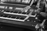 A vintage organ with dual keyboards and multiple buttons and switches on its console is partially obscured by large, glossy leaves of a nearby plant. Several VHS tapes or books are stacked horizontally on a shelf behind the organ.