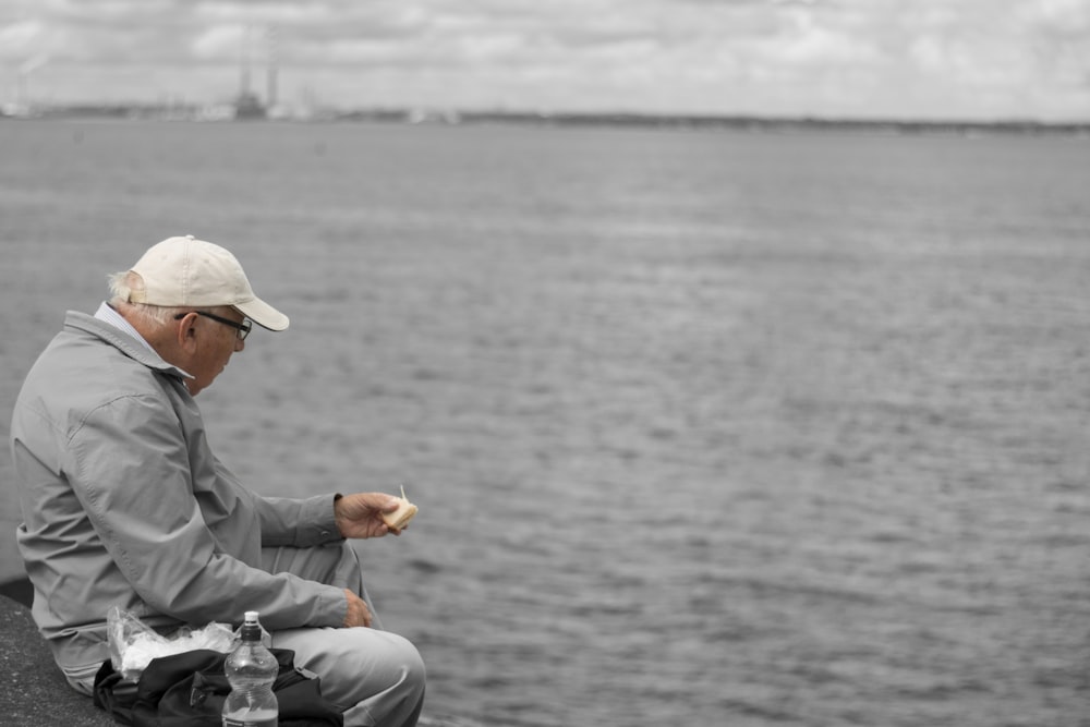 man in gray dress shirt and white pants sitting on gray rock near body of water