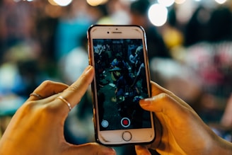 A pair of hands holding a smartphone, with a video being recorded on the screen. The background appears out of focus, featuring blurred lights and shapes, suggesting a busy environment such as a crowded market or event.