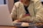 A person is sitting at a table working on a laptop. They are wearing a beige jacket and a dark shirt. On the table, there is a plate with a powdered dessert and a pair of glasses. The setting appears to be indoors, likely in a cafe or similar environment.