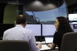 Two people are sitting in front of multiple computer monitors in a dimly lit room. One person is facing the monitors, while the other is turned slightly, appearing to engage in conversation or collaboration. The screens display various technical data, suggesting a work environment focused on analysis or monitoring.