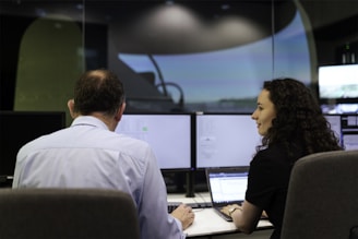 Two people are sitting in front of multiple computer monitors in a dimly lit room. One person is facing the monitors, while the other is turned slightly, appearing to engage in conversation or collaboration. The screens display various technical data, suggesting a work environment focused on analysis or monitoring.