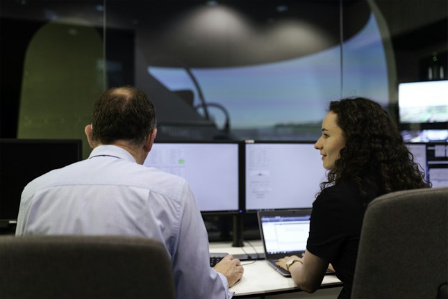 Two people are sitting in front of multiple computer monitors in a dimly lit room. One person is facing the monitors, while the other is turned slightly, appearing to engage in conversation or collaboration. The screens display various technical data, suggesting a work environment focused on analysis or monitoring.