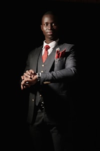 A man dressed in a sharp, dark suit with a red tie and matching pocket square stands with a confident posture. The suit jacket features gold buttons and there is a small floral pin on the lapel. The background is dark, creating a dramatic contrast that emphasizes the elegance of the attire. The lighting highlights the man's face and hands, giving a sense of sophistication and poise.