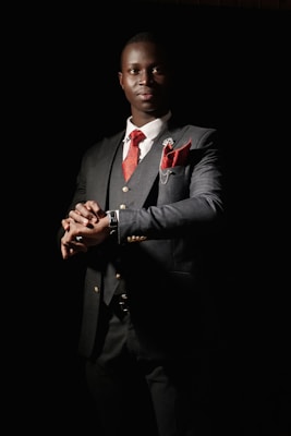 A man dressed in a sharp, dark suit with a red tie and matching pocket square stands with a confident posture. The suit jacket features gold buttons and there is a small floral pin on the lapel. The background is dark, creating a dramatic contrast that emphasizes the elegance of the attire. The lighting highlights the man's face and hands, giving a sense of sophistication and poise.