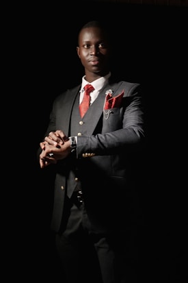 A man dressed in a sharp, dark suit with a red tie and matching pocket square stands with a confident posture. The suit jacket features gold buttons and there is a small floral pin on the lapel. The background is dark, creating a dramatic contrast that emphasizes the elegance of the attire. The lighting highlights the man's face and hands, giving a sense of sophistication and poise.