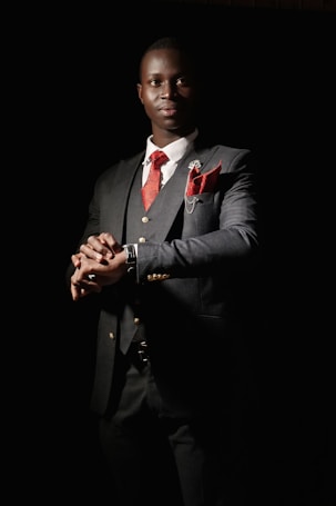 A man dressed in a sharp, dark suit with a red tie and matching pocket square stands with a confident posture. The suit jacket features gold buttons and there is a small floral pin on the lapel. The background is dark, creating a dramatic contrast that emphasizes the elegance of the attire. The lighting highlights the man's face and hands, giving a sense of sophistication and poise.