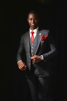 A well-dressed individual stands confidently against a dark background. The person is wearing a dark suit with a white shirt, a red tie, and a red pocket square. A decorative pin is attached to the suit's lapel. A watch is visible on the left wrist, adding a touch of elegance.