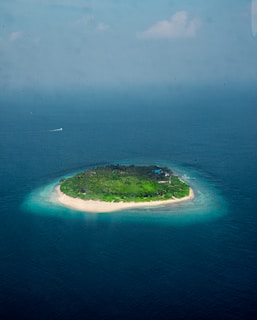 A small tropical island surrounded by turquoise waters. The island is covered in lush greenery with a sandy beach encircling it. A boat is visible near the island, and the vast ocean dominates the background under a slightly overcast sky.