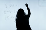 A person stands in front of a whiteboard writing mathematical equations. The silhouette is visible against the softly illuminated board filled with handwritten mathematical expressions.