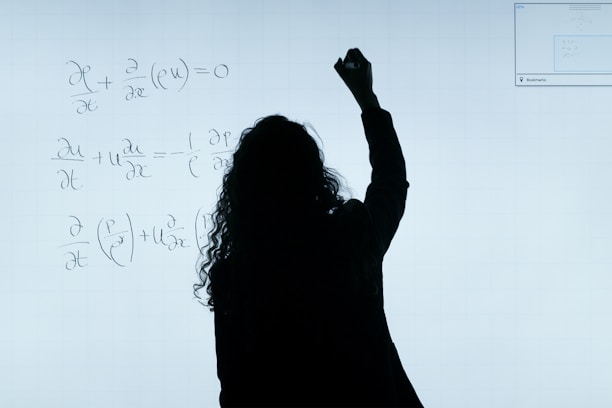 A person stands in front of a whiteboard writing mathematical equations. The silhouette is visible against the softly illuminated board filled with handwritten mathematical expressions.