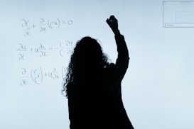 A person stands in front of a whiteboard writing mathematical equations. The silhouette is visible against the softly illuminated board filled with handwritten mathematical expressions.