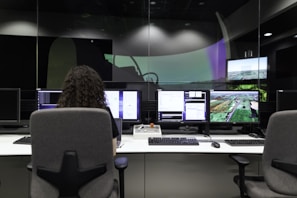 A person with curly hair is seated in front of multiple computer monitors displaying various information and images. The room appears to be a control or monitoring center, with dim lighting and specialized equipment. The atmosphere seems professional and focused.