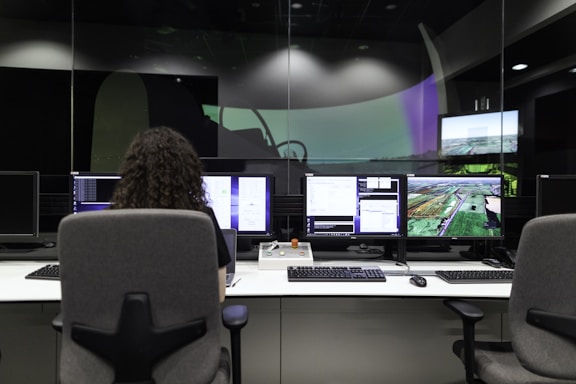 A person with curly hair is seated in front of multiple computer monitors displaying various information and images. The room appears to be a control or monitoring center, with dim lighting and specialized equipment. The atmosphere seems professional and focused.