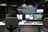 A person with curly hair is seated in front of multiple computer monitors displaying various information and images. The room appears to be a control or monitoring center, with dim lighting and specialized equipment. The atmosphere seems professional and focused.