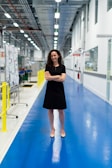 A woman with curly hair is standing confidently in the middle of a brightly lit industrial corridor. She is wearing a black dress and beige shoes, with her arms crossed and a friendly smile. The corridor is spacious with a blue glossy floor and white walls. Along the left side, there are bulletin boards and various industrial equipment.