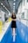 A woman with curly hair is standing confidently in the middle of a brightly lit industrial corridor. She is wearing a black dress and beige shoes, with her arms crossed and a friendly smile. The corridor is spacious with a blue glossy floor and white walls. Along the left side, there are bulletin boards and various industrial equipment.