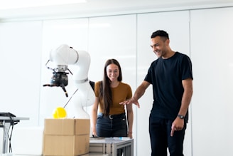 Two people, a woman and a man, are standing next to a robotic arm in a bright, modern room. The woman, wearing glasses and a brown top, is smiling while observing the robot. The man, dressed in a black t-shirt, is gesturing towards the robot with a cheerful expression. A cardboard box and other small objects are placed on the table in front of them.