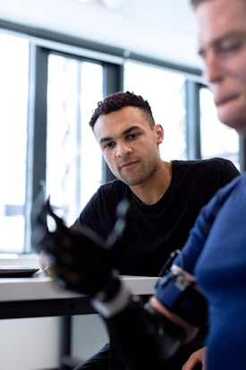A person wearing a black shirt is attentively observing a robotic hand. Another individual is partially visible in the foreground, seemingly interacting with the robotic device. The setting appears to be a brightly lit room with large windows in the background.