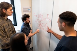A group of four people are engaged in a collaborative discussion around a whiteboard covered in mathematical equations and graphs drawn in red marker. They appear attentive and focused, with one person actively explaining or discussing the content on the board.