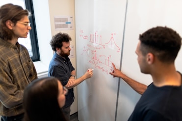 A group of four people are engaged in a collaborative discussion around a whiteboard covered in mathematical equations and graphs drawn in red marker. They appear attentive and focused, with one person actively explaining or discussing the content on the board.
