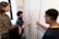 A group of four people are engaged in a collaborative discussion around a whiteboard covered in mathematical equations and graphs drawn in red marker. They appear attentive and focused, with one person actively explaining or discussing the content on the board.