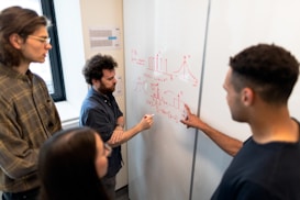 A group of four people are engaged in a collaborative discussion around a whiteboard covered in mathematical equations and graphs drawn in red marker. They appear attentive and focused, with one person actively explaining or discussing the content on the board.