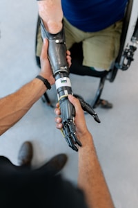 A close-up interaction between two individuals, focusing on one person fitting a prosthetic arm onto another's arm. The prosthetic is sleek and metallic, suggesting advanced technology. The scene suggests a sense of care and assistance.