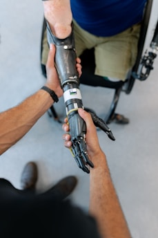 A close-up interaction between two individuals, focusing on one person fitting a prosthetic arm onto another's arm. The prosthetic is sleek and metallic, suggesting advanced technology. The scene suggests a sense of care and assistance.