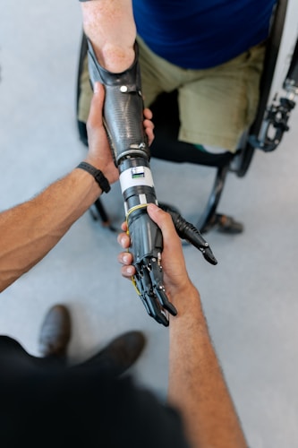 A close-up interaction between two individuals, focusing on one person fitting a prosthetic arm onto another's arm. The prosthetic is sleek and metallic, suggesting advanced technology. The scene suggests a sense of care and assistance.