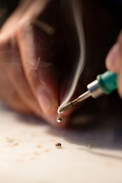 A hand holds a soldering iron that is melting a piece of solder, with visible smoke rising from the heated metal. The background is blurred, focusing attention on the soldering process and the small drop of molten solder.