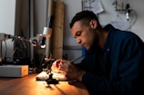 A person is focused on work at a desk equipped with a microscope and other small tools, suggesting a detailed task such as electronics repair or scientific research. The environment is dimly lit, emphasizing the concentration and meticulousness of the activity.
