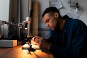 A person is focused on work at a desk equipped with a microscope and other small tools, suggesting a detailed task such as electronics repair or scientific research. The environment is dimly lit, emphasizing the concentration and meticulousness of the activity.