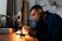 A person is focused on work at a desk equipped with a microscope and other small tools, suggesting a detailed task such as electronics repair or scientific research. The environment is dimly lit, emphasizing the concentration and meticulousness of the activity.