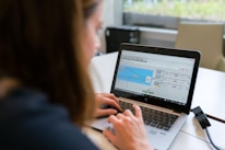 A person is working on a laptop at a table, focusing on a software interface displayed on the screen. The interface appears technical or related to programming and data flow. The setting seems to be a modern indoor office or study area with a window in the background.