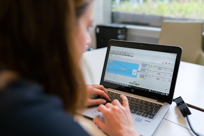 A person is working on a laptop at a table, focusing on a software interface displayed on the screen. The interface appears technical or related to programming and data flow. The setting seems to be a modern indoor office or study area with a window in the background.