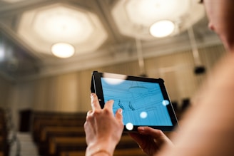 A person is holding a tablet with a digital drawing displayed on it. The setting appears to be an auditorium with empty seating in the background and a bright, well-lit ceiling above. The person is likely using the tablet to create or view architectural designs.