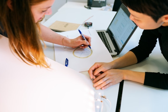 Two people are collaborating at a desk. One person is drawing on a sheet of paper with a pen, while the other is observing. A laptop is open nearby, displaying a document. Various office supplies, including notebooks and a calculator, are scattered on the desk.