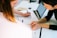 Two people are collaborating at a desk. One person is drawing on a sheet of paper with a pen, while the other is observing. A laptop is open nearby, displaying a document. Various office supplies, including notebooks and a calculator, are scattered on the desk.