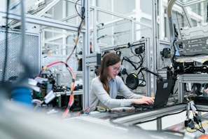 A woman is working on a laptop surrounded by various electronic and mechanical equipment in a laboratory or industrial setting. The environment appears to be focused on technology with visible complex wiring and machinery.