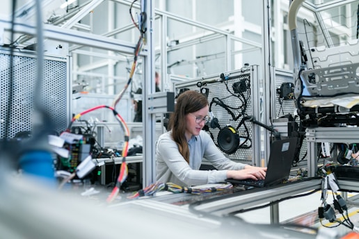 A woman is working on a laptop surrounded by various electronic and mechanical equipment in a laboratory or industrial setting. The environment appears to be focused on technology with visible complex wiring and machinery.