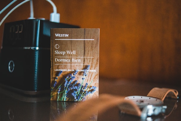 A small card from Westin with the text 'Sleep Well' and 'Dormez Bien' is placed on a surface. It features an image of lavender and mentions lavender balm. Next to it is an electronic device with a digital clock display, and in the foreground, part of a wristwatch with a brown leather strap is visible.