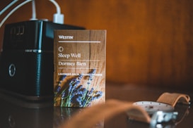 A small card from Westin with the text 'Sleep Well' and 'Dormez Bien' is placed on a surface. It features an image of lavender and mentions lavender balm. Next to it is an electronic device with a digital clock display, and in the foreground, part of a wristwatch with a brown leather strap is visible.