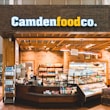 A small convenience store or food shop features a variety of snacks, drinks, and quick meals displayed on shelves, with a counter for service. The store has a wooden and brick design, with warm lighting hanging from the ceiling. Products like bottled water, chips, and packaged goods are visible.