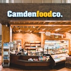A small convenience store or food shop features a variety of snacks, drinks, and quick meals displayed on shelves, with a counter for service. The store has a wooden and brick design, with warm lighting hanging from the ceiling. Products like bottled water, chips, and packaged goods are visible.
