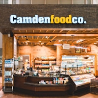 A small convenience store or food shop features a variety of snacks, drinks, and quick meals displayed on shelves, with a counter for service. The store has a wooden and brick design, with warm lighting hanging from the ceiling. Products like bottled water, chips, and packaged goods are visible.