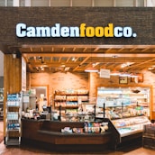 A small convenience store or food shop features a variety of snacks, drinks, and quick meals displayed on shelves, with a counter for service. The store has a wooden and brick design, with warm lighting hanging from the ceiling. Products like bottled water, chips, and packaged goods are visible.
